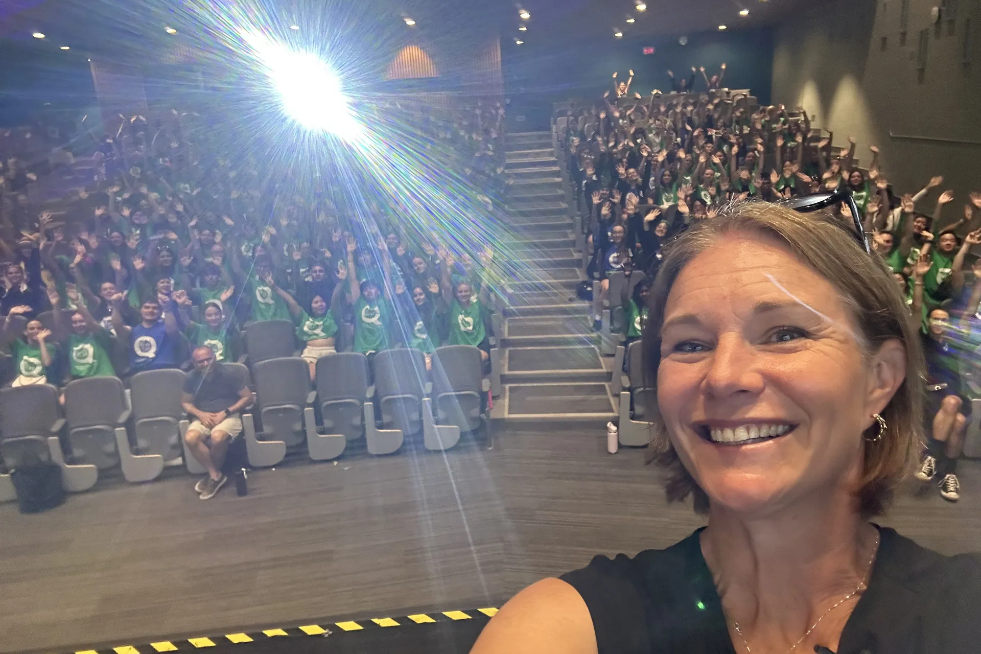 Kim Lockhart taking a selfie on stage in front of a packed lecture hall of students waving their arms.